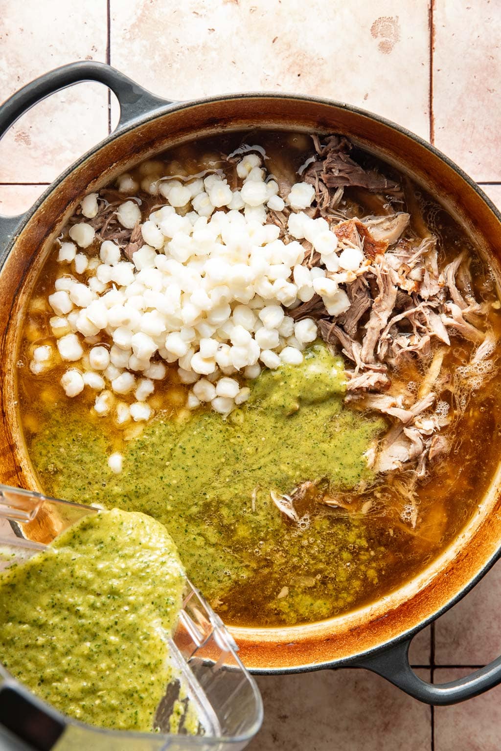 Shredded pork, green sauce, and hominy being added to the stock in a dutch oven