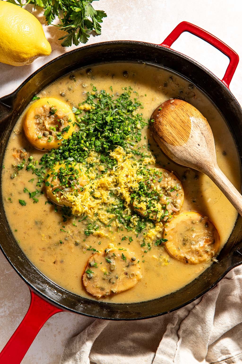 herbs and lemon zest being added to piccata sauce in a skillet