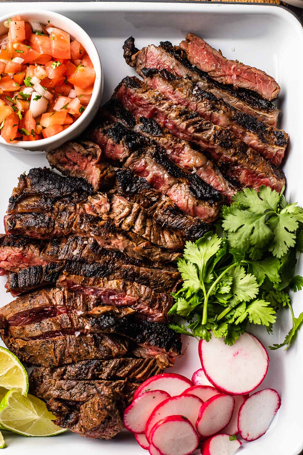 Sliced carne asada on baking tray with radish, cilantro, lime, and pico de gallo. 