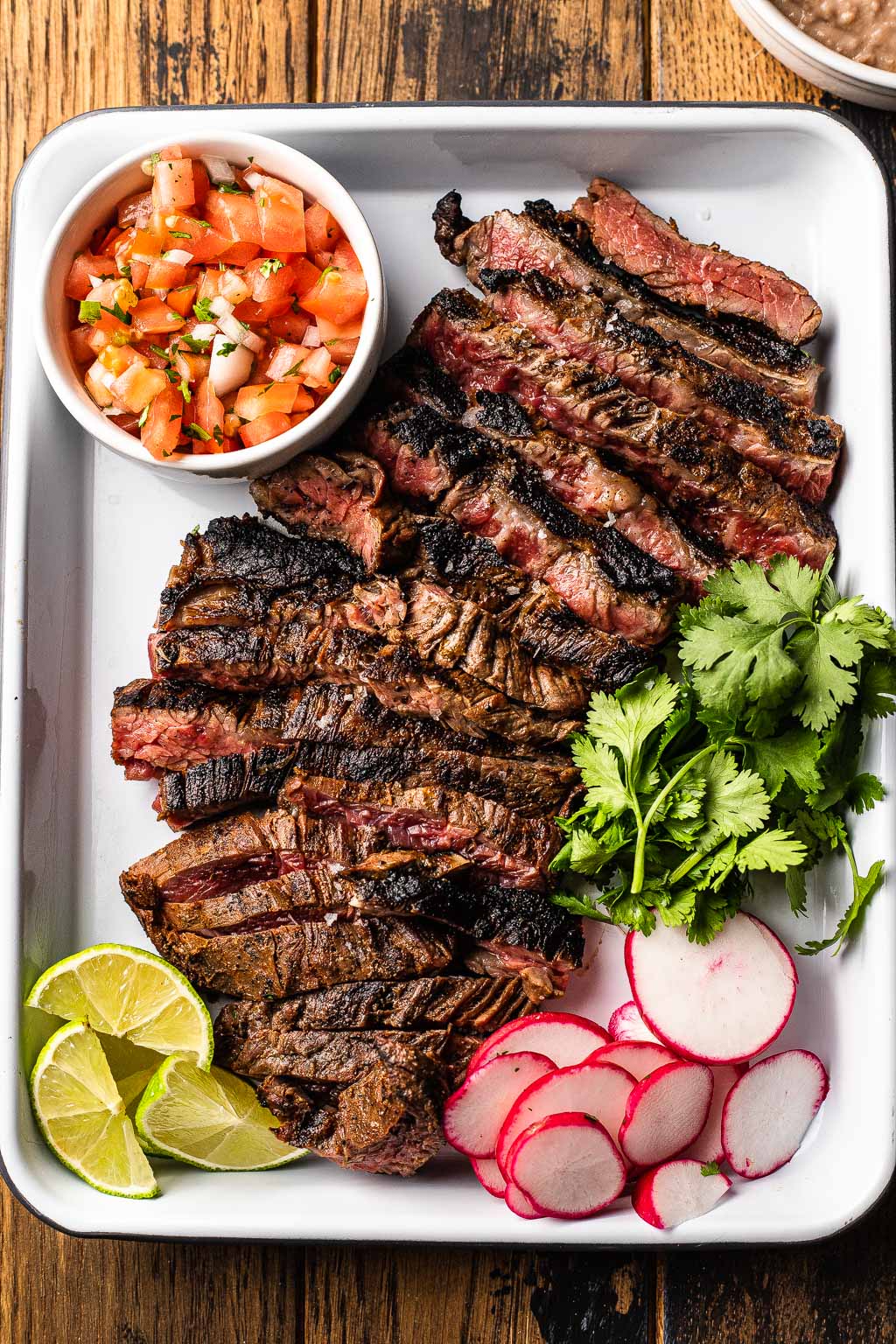 Sliced carne asada on baking tray with radish, cilantro, lime, and pico de gallo. 