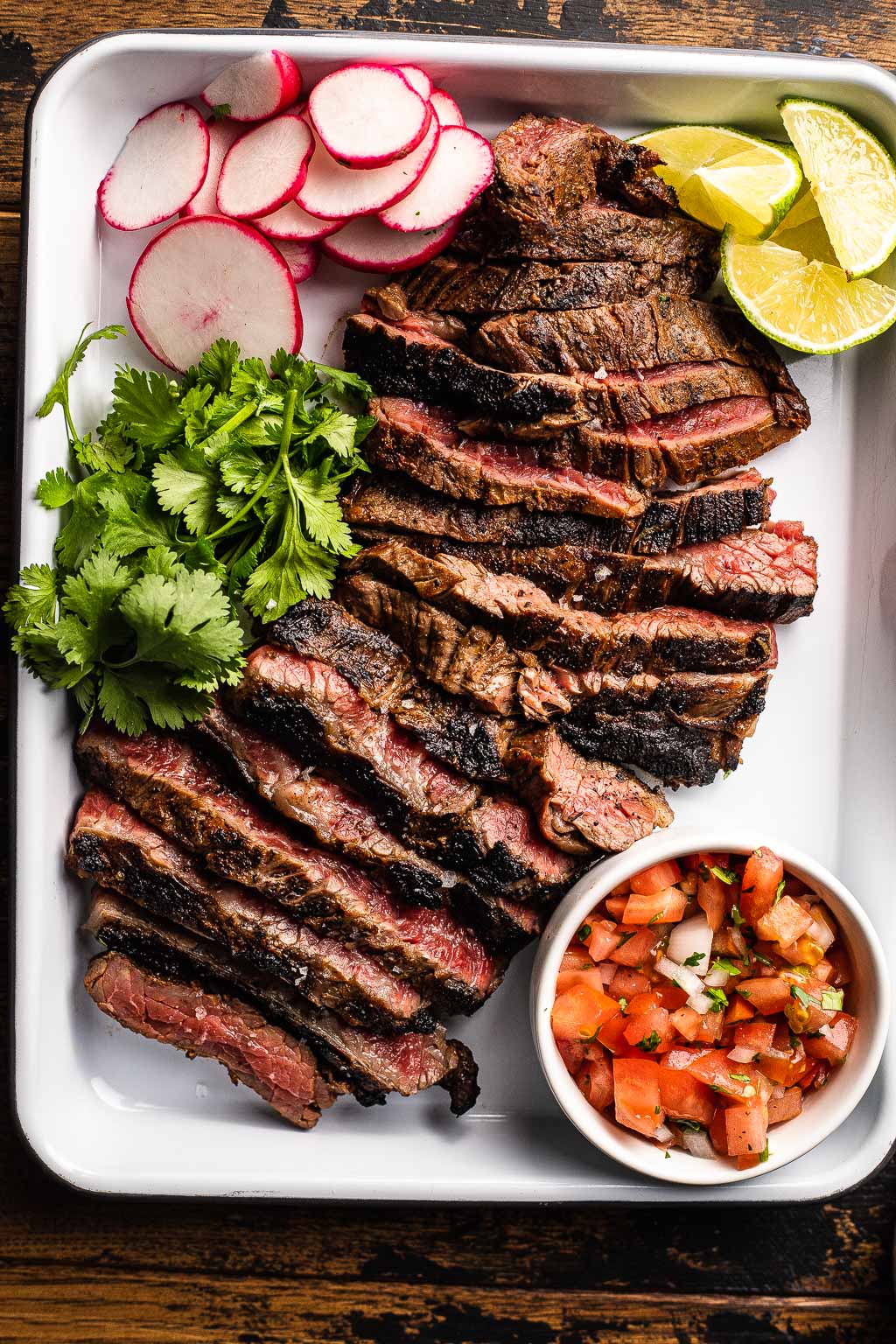 Sliced carne asada on baking tray with radish, cilantro, lime, and pico de gallo. 